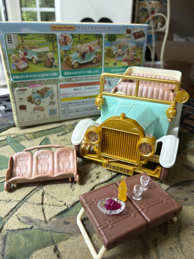 May include: A toy set featuring a vintage-style car in light blue and gold, with a matching bench and picnic table. Accessories include a bottle, glasses, and a plate of grapes. The box reads "Sylvanian Families".