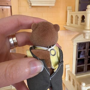 May include: A brown bear figurine wearing a yellow shirt and a dark gray vest with a silver ring detail. The bear is part of a miniature dollhouse set, with a yellow and white building in the background.