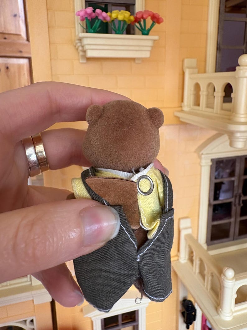 May include: A brown bear figurine wearing a yellow shirt and a dark gray vest with a silver ring detail. The bear is part of a miniature dollhouse set, with a yellow and white building in the background.