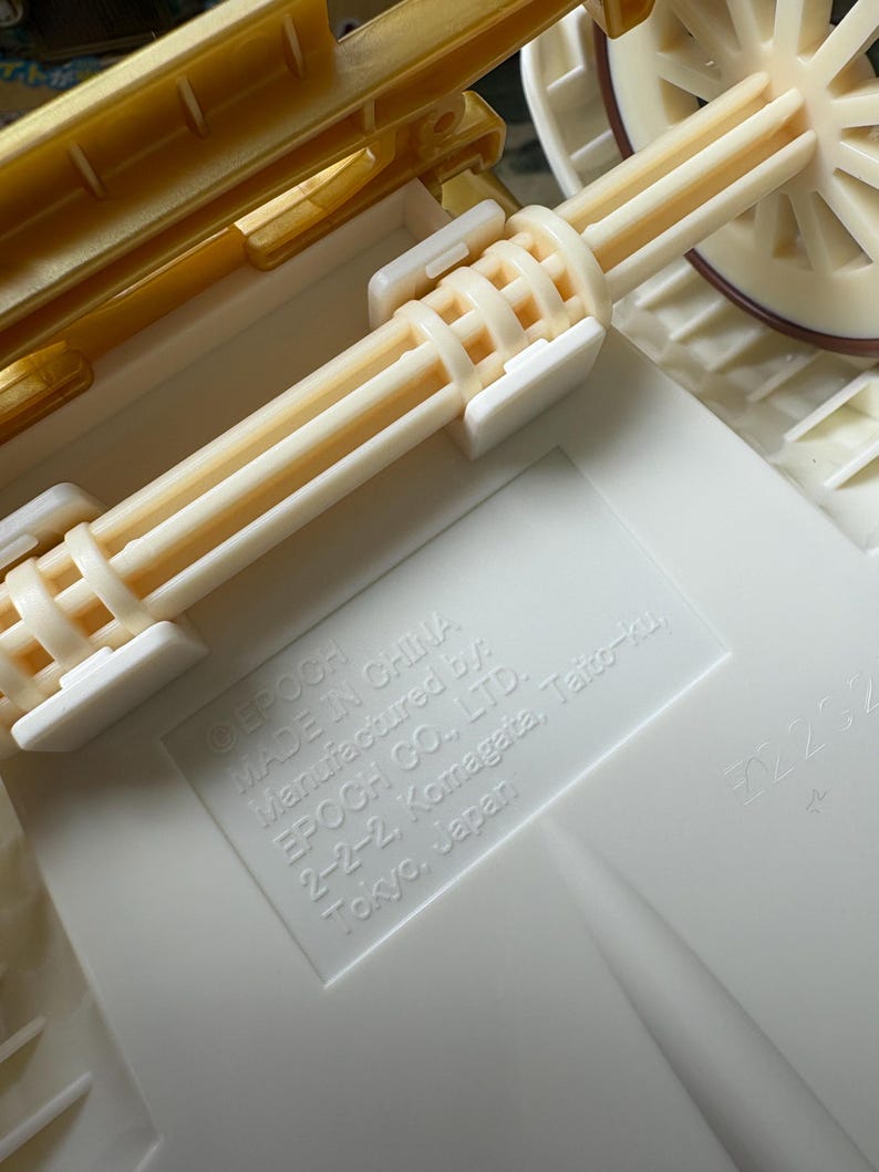 May include: Close-up of a toy carriage with a cream-colored body and wheels. The carriage has a gold-colored top and cream-colored axles. The text "MADE IN CHINA" and "EPOCH CO., LTD." are visible.
