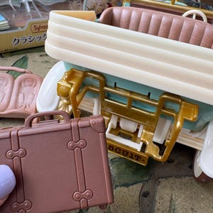 May include: A miniature brown suitcase with a handle and decorative clasps is held near a toy car. The car is cream, gold, and teal, with a brown interior. The toy car has a vintage aesthetic.