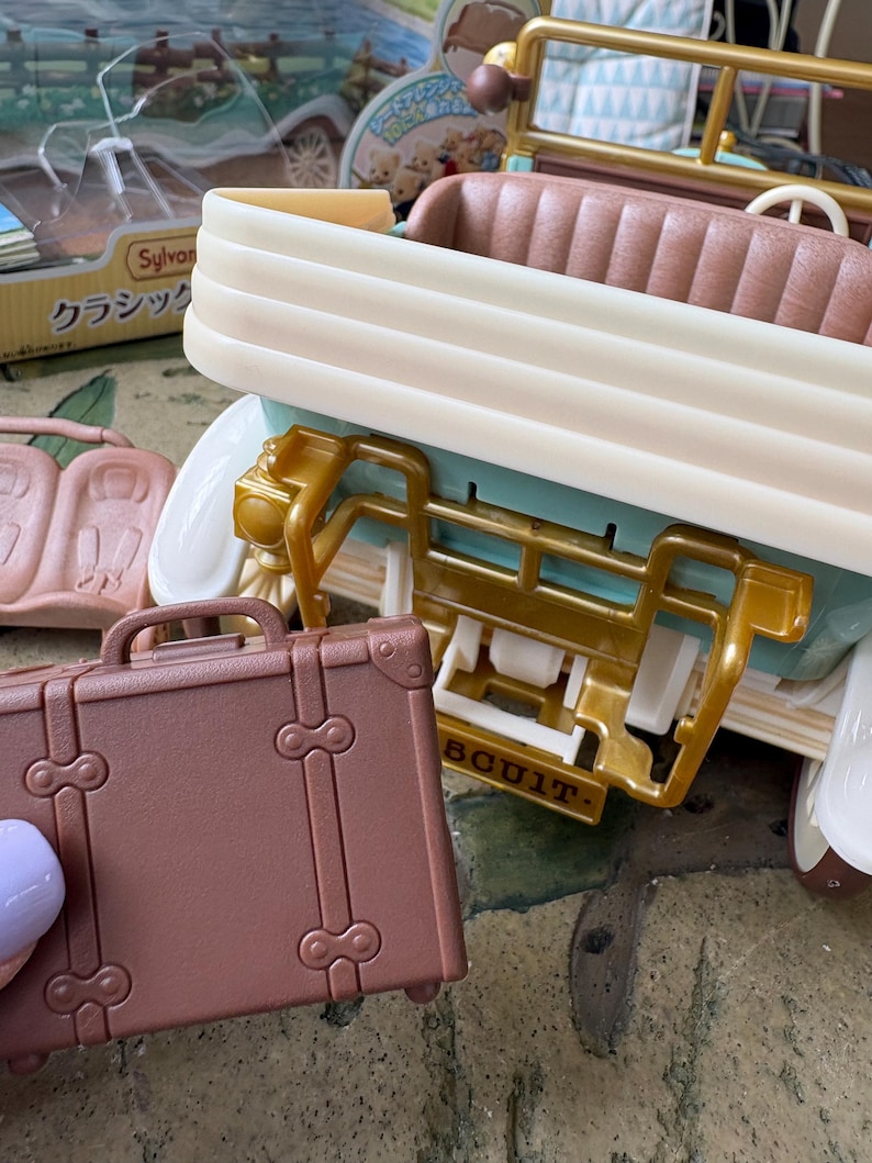 May include: A miniature brown suitcase with a handle and decorative clasps is held near a toy car. The car is cream, gold, and teal, with a brown interior. The toy car has a vintage aesthetic.
