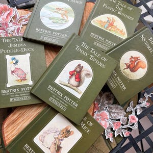 May include: A collection of Beatrix Potter books with green covers and illustrations. Titles include "The Tale of Mr. Jeremy Fisher," "The Tale of the Flopsy Bunnies," and "The Tale of Timmy Tiptoes." The books are arranged on a wooden surface with floral decorations.