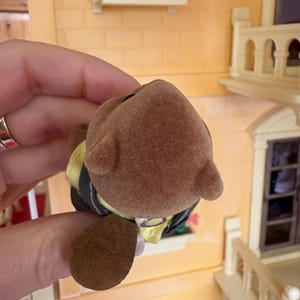 May include: A brown toy bear with a yellow and black outfit. The bear has a fuzzy texture and small ears. The toy is held in front of a miniature house with a yellow exterior and decorative flower boxes.