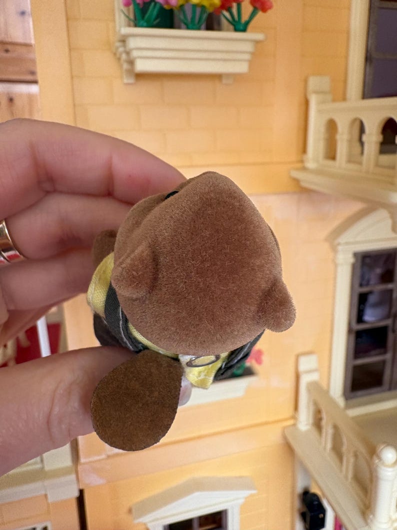 May include: A brown toy bear with a yellow and black outfit. The bear has a fuzzy texture and small ears. The toy is held in front of a miniature house with a yellow exterior and decorative flower boxes.