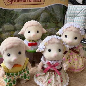 May include: Four Sylvanian Families sheep figures, each dressed in different outfits. The sheep have cream-coloured fur and pink noses. One wears a yellow top and green patterned shorts. The box in the background reads "Sylvanian Families".