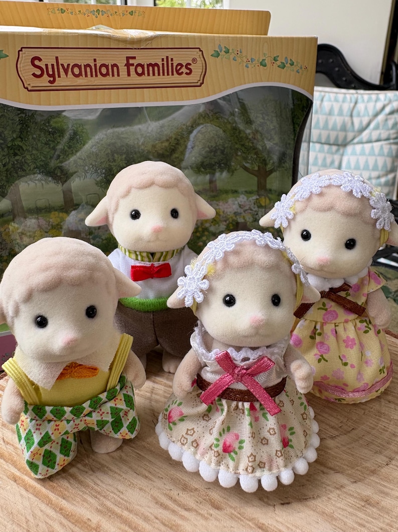 May include: Four Sylvanian Families sheep figures, each dressed in different outfits. The sheep have cream-coloured fur and pink noses. One wears a yellow top and green patterned shorts. The box in the background reads "Sylvanian Families".