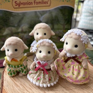 May include: Four Sylvanian Families sheep figures. The sheep are wearing different outfits, including dresses and dungarees. The figures are displayed on a wooden surface, with the Sylvanian Families box visible.