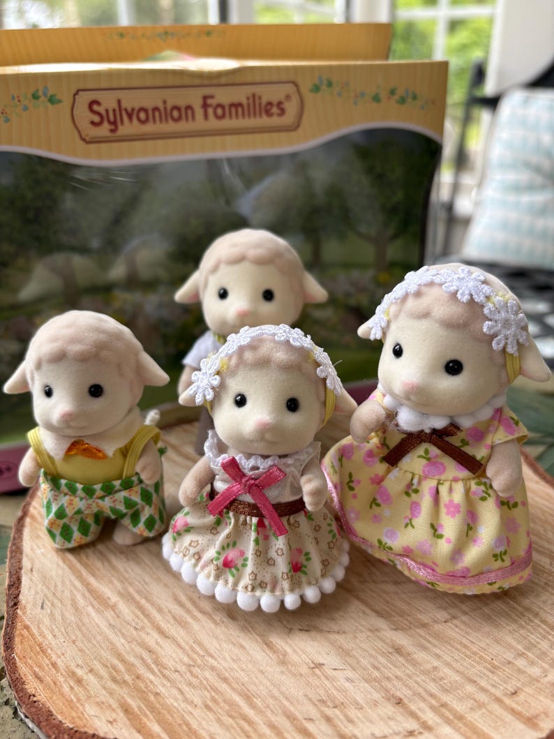 May include: Four Sylvanian Families sheep figures. The sheep are wearing different outfits, including dresses and dungarees. The figures are displayed on a wooden surface, with the Sylvanian Families box visible.