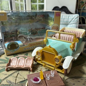 May include: A toy car set featuring a light blue vintage car with gold accents, white fenders, and a tan interior. Includes a brown bench, a brown table with miniature food and drinks, and a box with Japanese text.
