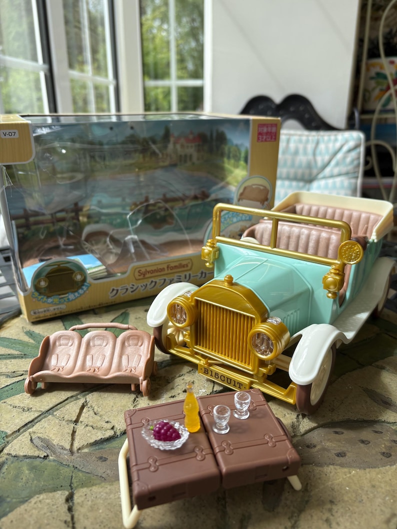 May include: A toy car set featuring a light blue vintage car with gold accents, white fenders, and a tan interior. Includes a brown bench, a brown table with miniature food and drinks, and a box with Japanese text.