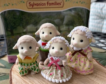 May include: A Sylvanian Families sheep family set featuring four flocked animal figures. The figures include two adults and two children dressed in colorful patterned clothing, with the children wearing white floral headbands, displayed in front of their original box.