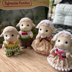May include: Four Sylvanian Families sheep figures, each dressed in different outfits. One wears a green and yellow outfit, another a bow tie and vest, and two wear floral dresses. The figures are displayed on a wooden surface.