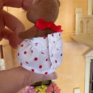 May include: A brown teddy bear figurine wearing a red bow and a white dress with red polka dots. The dress has a lace trim. The bear is part of a dollhouse set, with a miniature building and flower boxes in the background.