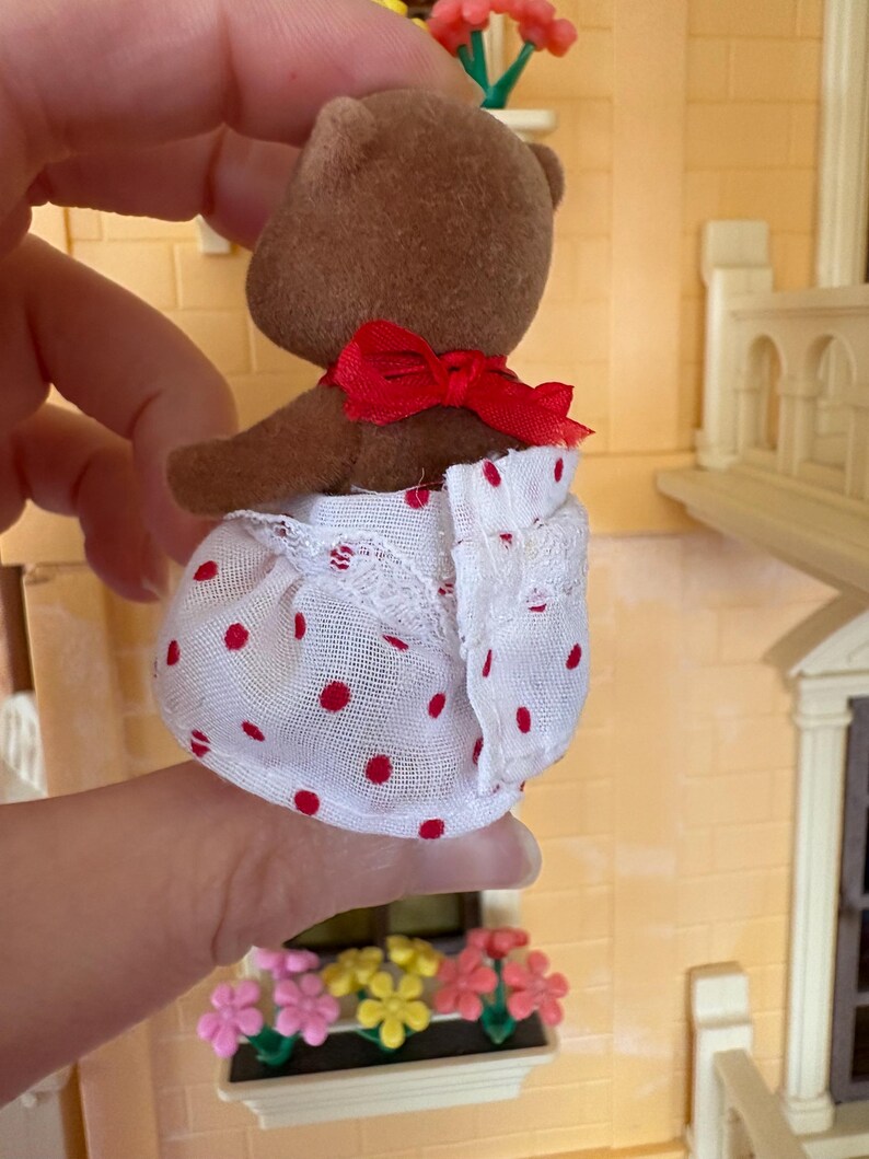 May include: A brown teddy bear figurine wearing a red bow and a white dress with red polka dots. The dress has a lace trim. The bear is part of a dollhouse set, with a miniature building and flower boxes in the background.