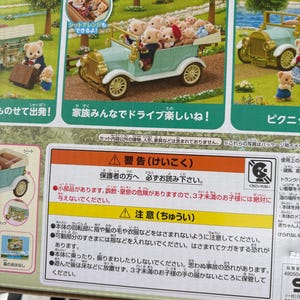 May include: A Sylvanian Families toy set featuring a light blue classic family car filled with bear figurines. The box also shows a picnic scene and a bear with luggage. The text says "Family fun drive!"