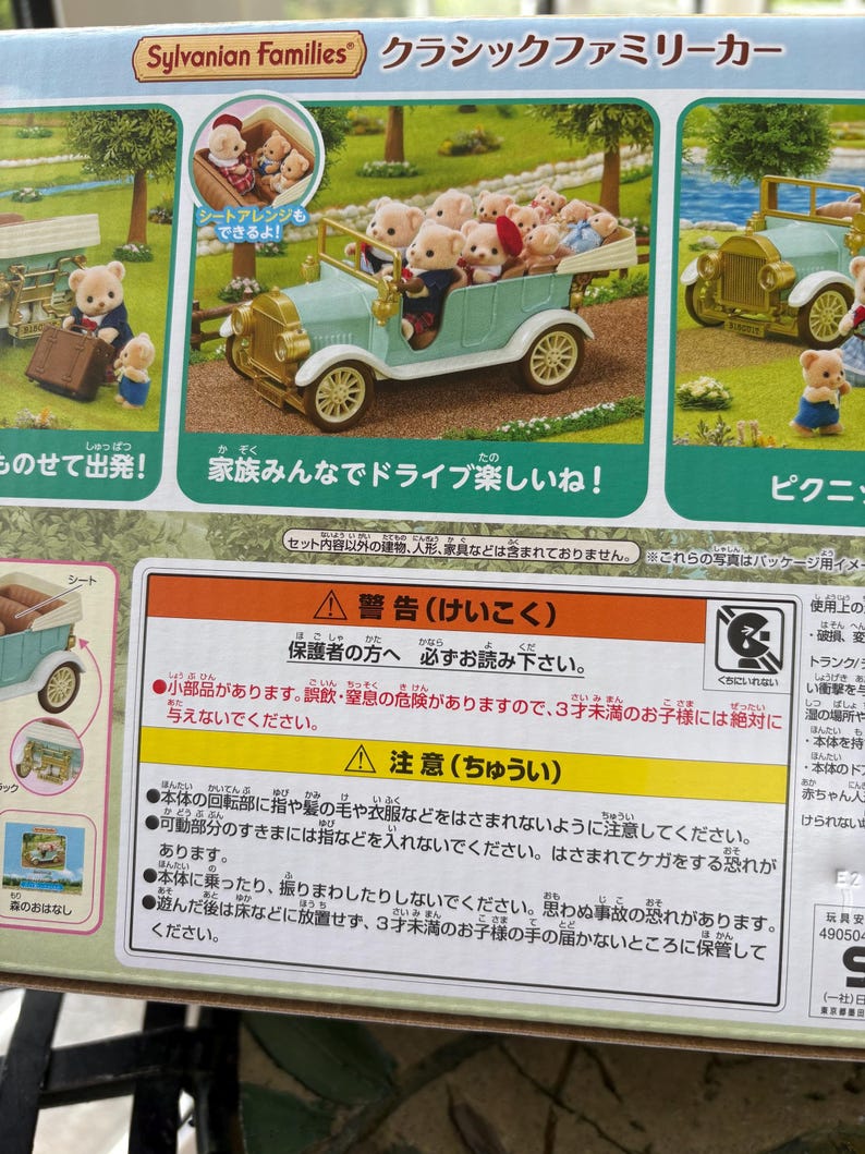 May include: A Sylvanian Families toy set featuring a light blue classic family car filled with bear figurines. The box also shows a picnic scene and a bear with luggage. The text says "Family fun drive!"