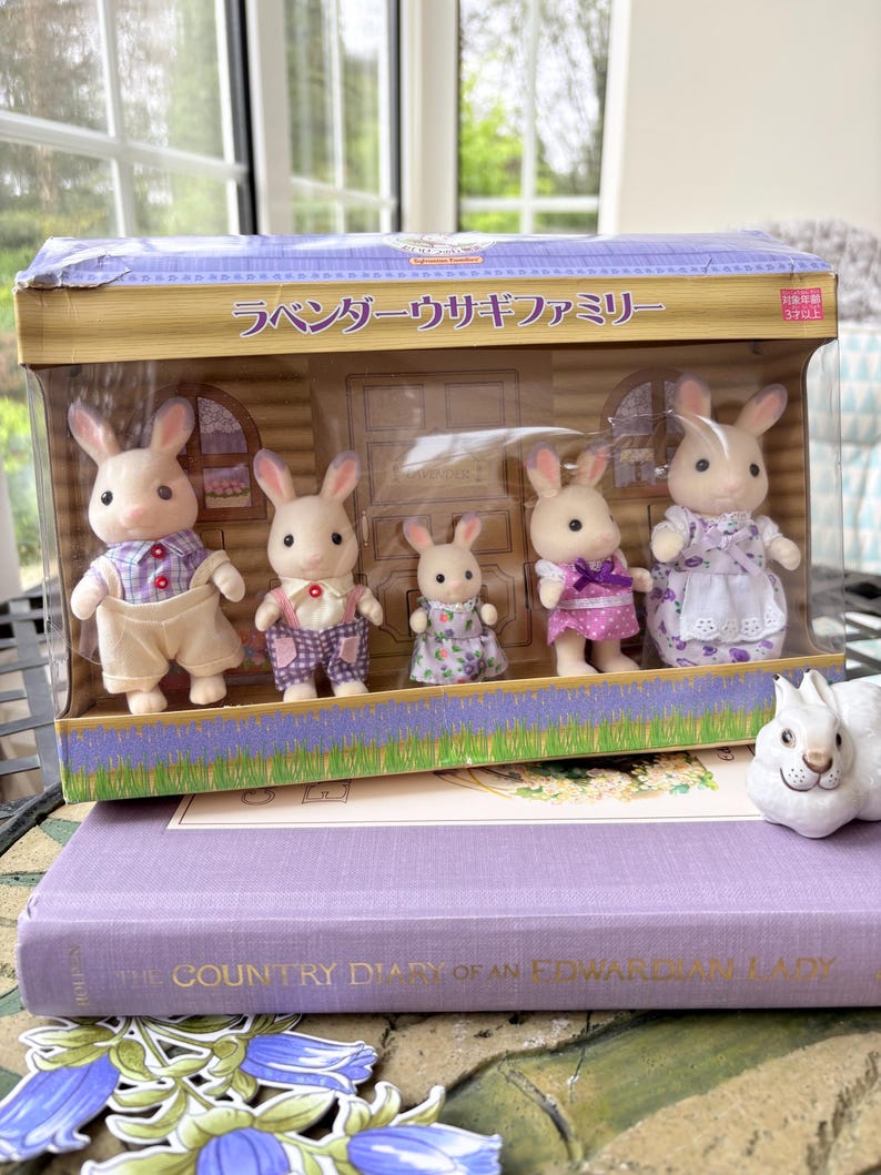 May include: A boxed set of toy rabbit family figures. The rabbits are dressed in various outfits, including overalls, dresses, and shirts. The box has Japanese text and a lavender and floral design. The set is displayed on a purple book.