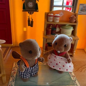 May include: Two brown beaver figurines, one wearing blue and white checkered overalls with orange straps, the other in a white skirt with red polka dots. A miniature kitchen scene with a clock and shelves is in the background.