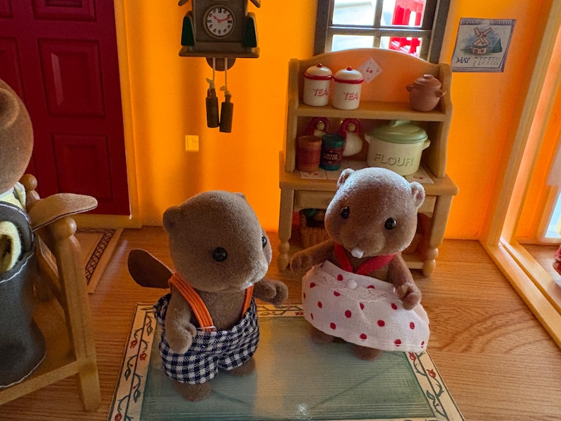 May include: Two brown beaver figurines, one wearing blue and white checkered overalls with orange straps, the other in a white skirt with red polka dots. A miniature kitchen scene with a clock and shelves is in the background.