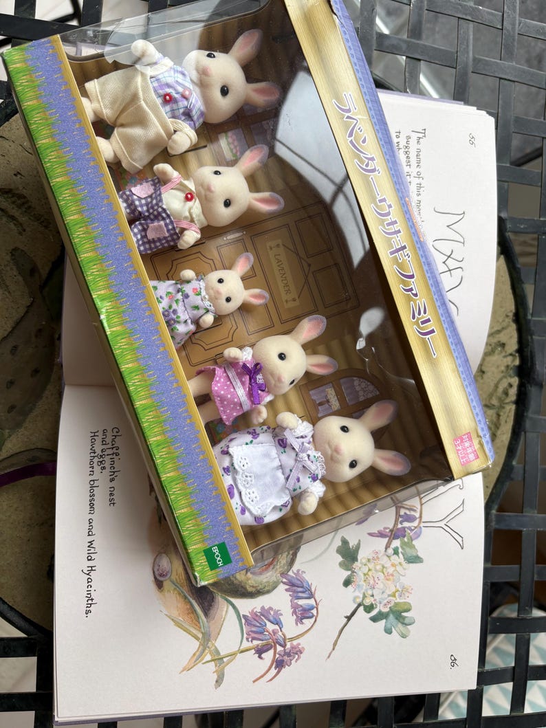 May include: A boxed set of toy rabbit figurines, each with a white body and dressed in various pastel-colored outfits. The box has Japanese text and is resting on an open book with illustrations of flowers and text.