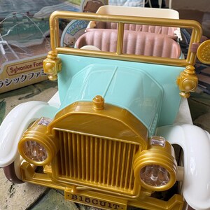 May include: A toy car in pastel colors, featuring a gold-colored front grill and headlights, with the word "BISCUIT" on the front. The car has a light blue body and white fenders. The interior is a light brown color.