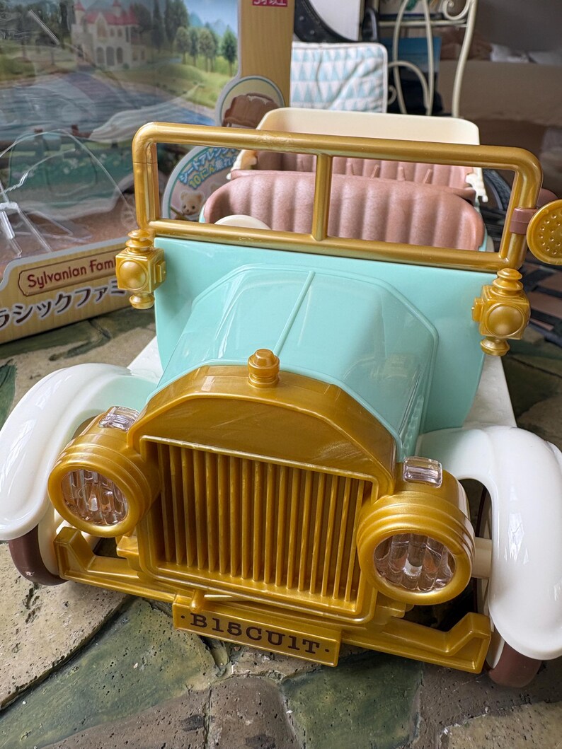 May include: A toy car in pastel colors, featuring a gold-colored front grill and headlights, with the word "BISCUIT" on the front. The car has a light blue body and white fenders. The interior is a light brown color.