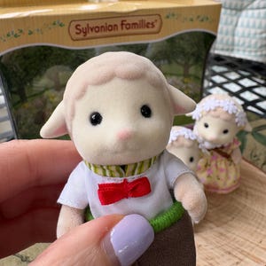 May include: A Sylvanian Families toy lamb figurine wearing a white shirt with a red bow tie, brown trousers, and a green and white striped collar. The toy is held in front of a box with the brand name.