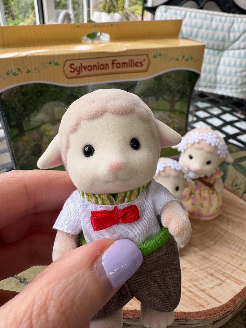May include: A Sylvanian Families toy lamb figurine wearing a white shirt with a red bow tie, brown trousers, and a green and white striped collar. The toy is held in front of a box with the brand name.