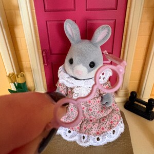 Może przedstawiać: Szara figurka kr&oacute;lika w r&oacute;żowej sukience w kwiaty z białym koronkowym wykończeniem. Kr&oacute;lik stoi przed r&oacute;żowymi drzwiami. Para r&oacute;żowych okular&oacute;w zabawkowych jest trzymana przed kr&oacute;likiem.