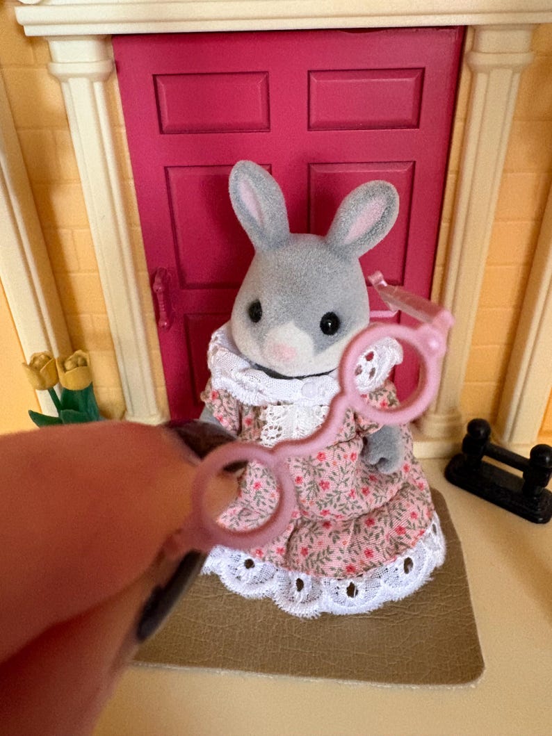 Może przedstawiać: Szara figurka kr&oacute;lika w r&oacute;żowej sukience w kwiaty z białym koronkowym wykończeniem. Kr&oacute;lik stoi przed r&oacute;żowymi drzwiami. Para r&oacute;żowych okular&oacute;w zabawkowych jest trzymana przed kr&oacute;likiem.