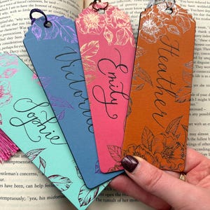 Può includere: Quattro segnalibri colorati con disegni floreali e nomi in corsivo: Sophie, Victoria, Emily e Heather. Ogni segnalibro ha un colore diverso, tra cui turchese, blu, rosa e arancione.