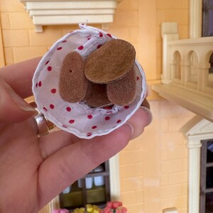 May include: A small, white fabric cone with red polka dots holds several brown, oval-shaped objects. The cone is held in front of a miniature building with window boxes filled with colorful flowers.
