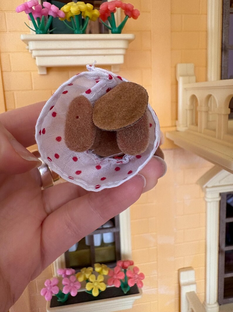 May include: A small, white fabric cone with red polka dots holds several brown, oval-shaped objects. The cone is held in front of a miniature building with window boxes filled with colorful flowers.