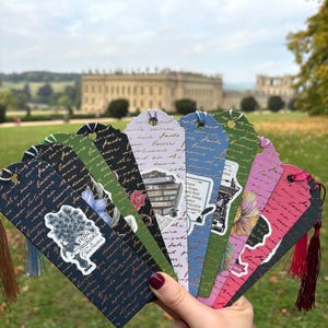May include: A selection of decorative bookmarks with tassel details, showcasing literary themes and quotes. Each bookmark features distinct designs, including floral illustrations, book stacks, and script lettering. The bookmarks are held in front of a blurred outdoor background.