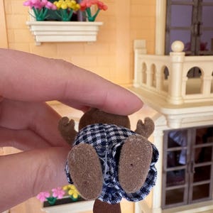 May include: A small, brown beaver figurine wearing a blue and white checkered outfit. The beaver has brown felt paws and a brown tail. The figurine is held up in front of a miniature house with a window box of flowers.