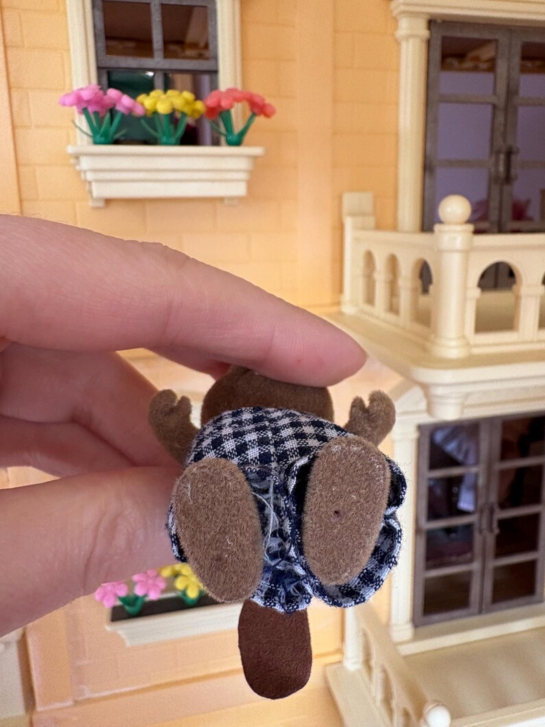 May include: A small, brown beaver figurine wearing a blue and white checkered outfit. The beaver has brown felt paws and a brown tail. The figurine is held up in front of a miniature house with a window box of flowers.
