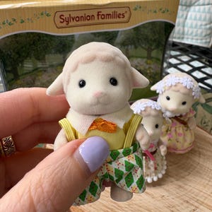 May include: A Sylvanian Families lamb figure in a yellow top, orange bow tie, and green patterned shorts. Two other lamb figures are visible, one in a floral dress and the other in a white dress. The box in the background has the words "Sylvanian Families".