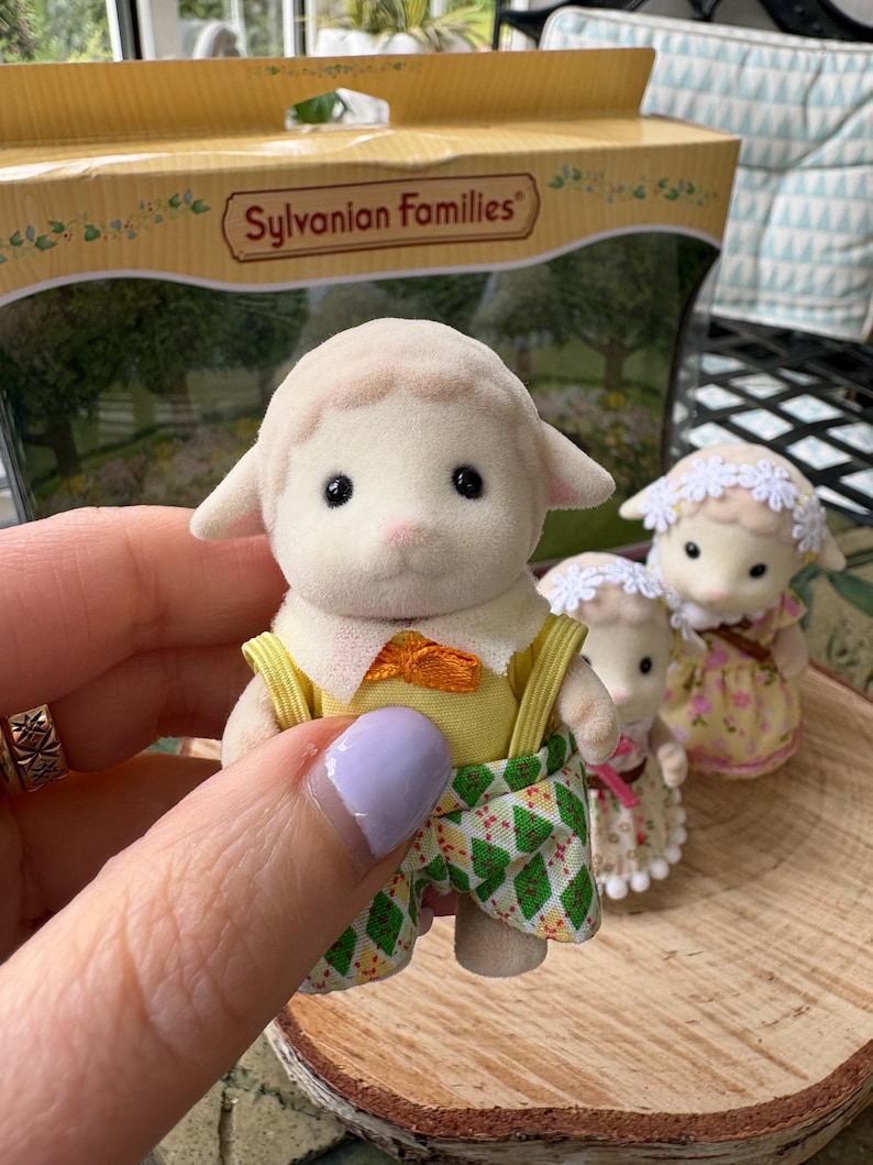 May include: A Sylvanian Families lamb figure in a yellow top, orange bow tie, and green patterned shorts. Two other lamb figures are visible, one in a floral dress and the other in a white dress. The box in the background has the words "Sylvanian Families".