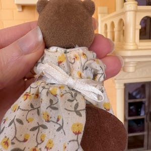May include: A brown bear figurine wearing a white dress with a yellow floral pattern. The dress has short sleeves and a white bow in the back. The bear has a brown tail and small ears.