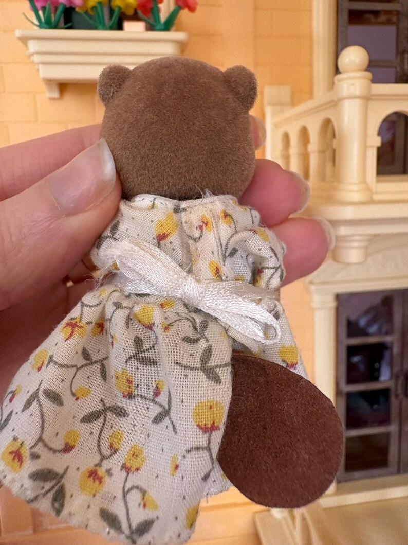 May include: A brown bear figurine wearing a white dress with a yellow floral pattern. The dress has short sleeves and a white bow in the back. The bear has a brown tail and small ears.