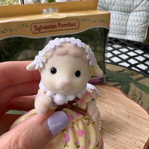 May include: A Sylvanian Families lamb figurine, dressed in a yellow floral dress and a white lace bonnet. The toy lamb is held in a hand, with a box in the background. The box has the words "Sylvanian Families" printed on it.