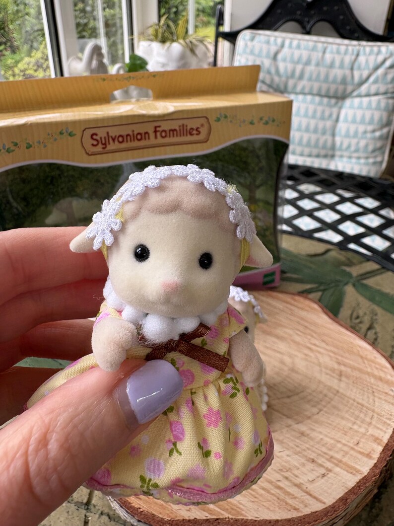May include: A Sylvanian Families lamb figurine, dressed in a yellow floral dress and a white lace bonnet. The toy lamb is held in a hand, with a box in the background. The box has the words "Sylvanian Families" printed on it.