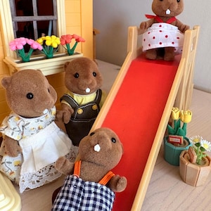 May include: A group of brown beaver figurines in various outfits. One beaver is at the top of a red slide, another is at the bottom, and two others are standing nearby. A yellow house with a window box of flowers is in the background.
