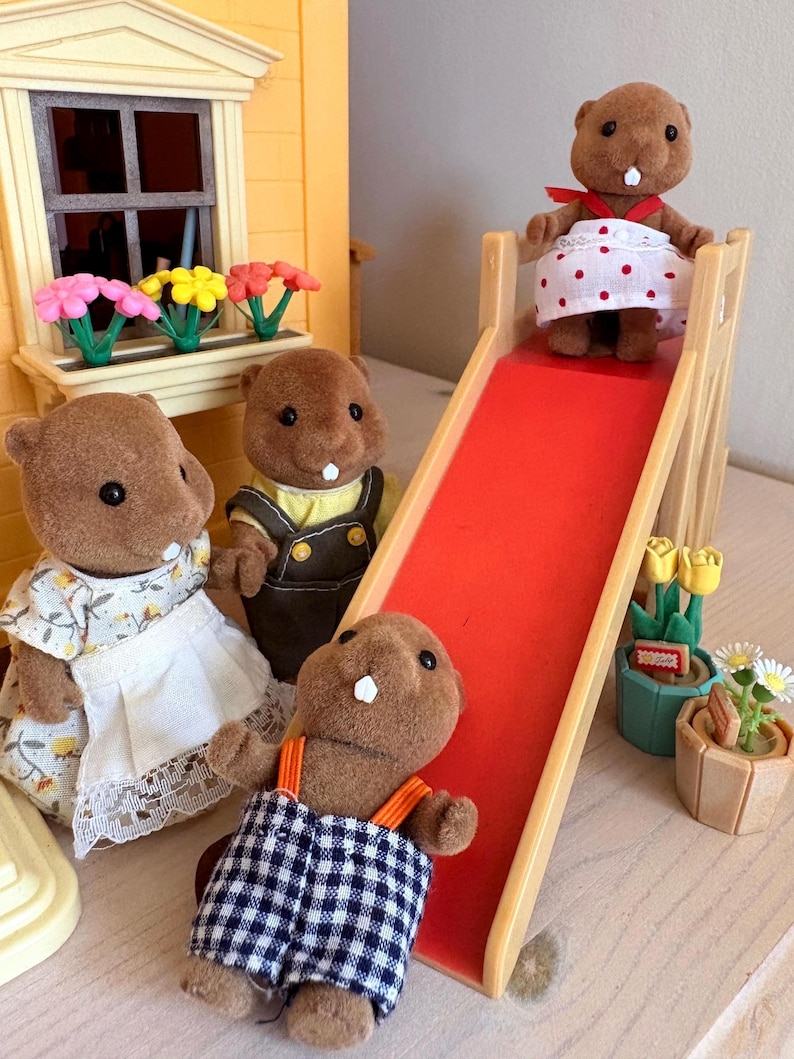 May include: A group of brown beaver figurines in various outfits. One beaver is at the top of a red slide, another is at the bottom, and two others are standing nearby. A yellow house with a window box of flowers is in the background.