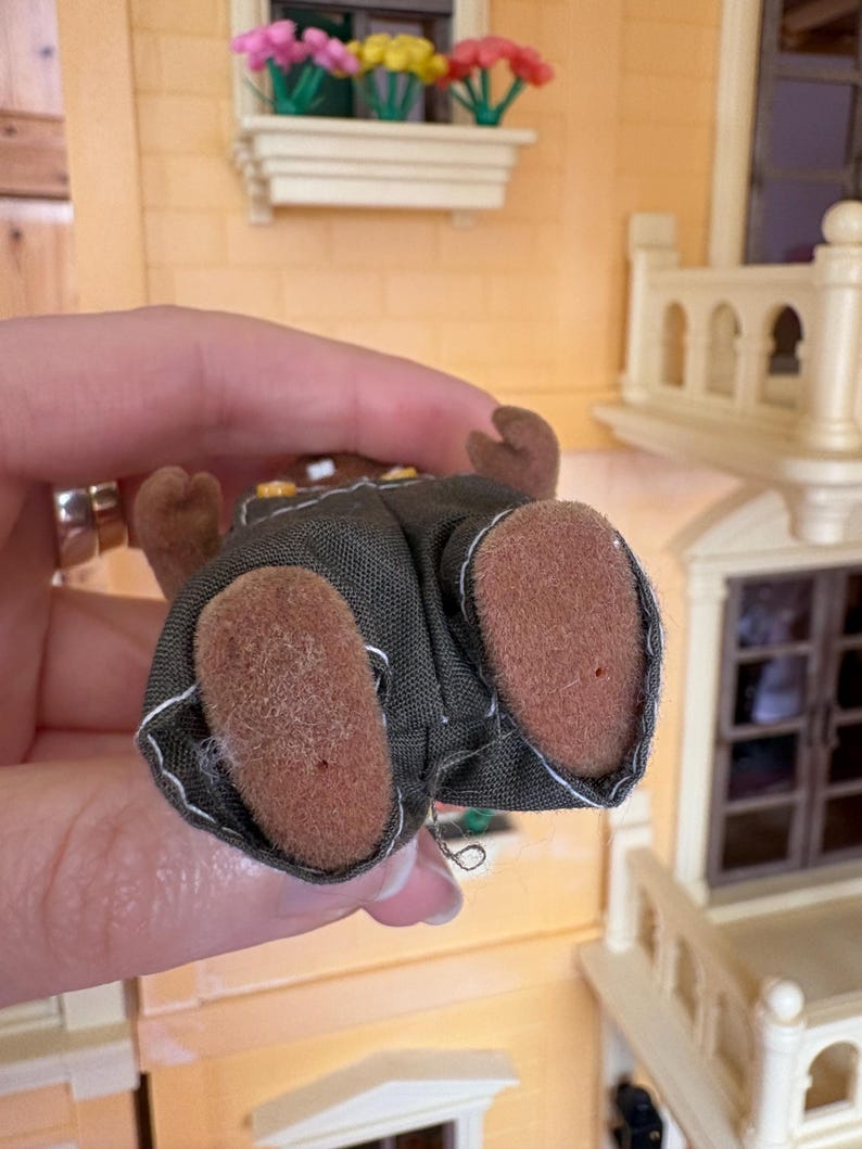 May include: A miniature brown bear figure wearing a dark green outfit with white stitching. The bear's brown feet are visible, and the figure is held against a backdrop of a dollhouse with a window box of flowers.