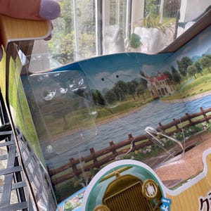 May include: A partially open toy box with a clear plastic window. The box features a scenic illustration of a river, trees, and a house. A vintage car is visible at the bottom. The box is made of cardboard and has Japanese text.