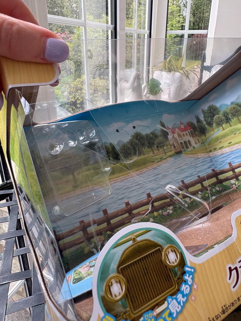 May include: A partially open toy box with a clear plastic window. The box features a scenic illustration of a river, trees, and a house. A vintage car is visible at the bottom. The box is made of cardboard and has Japanese text.