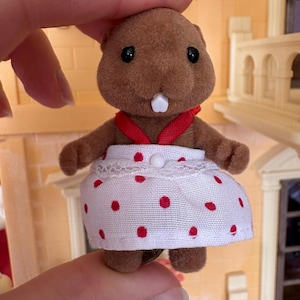 May include: A brown beaver figurine wearing a white dress with red polka dots and a red ribbon. The toy beaver has black eyes and a white tooth. The figurine is part of a dollhouse set.