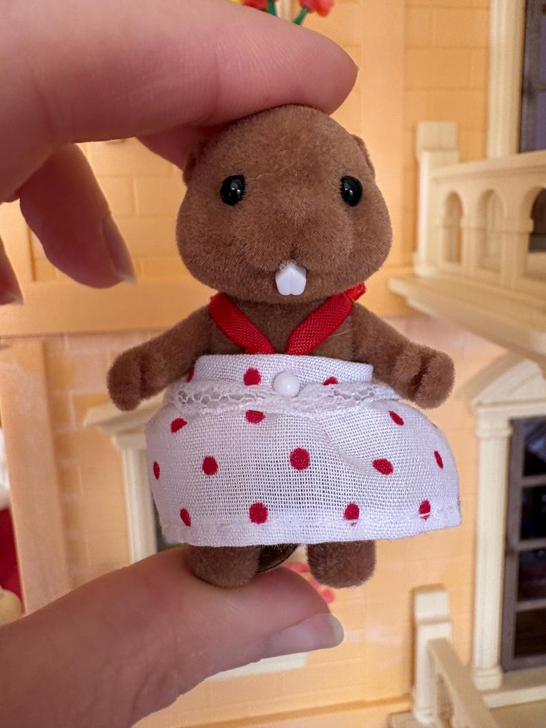 May include: A brown beaver figurine wearing a white dress with red polka dots and a red ribbon. The toy beaver has black eyes and a white tooth. The figurine is part of a dollhouse set.